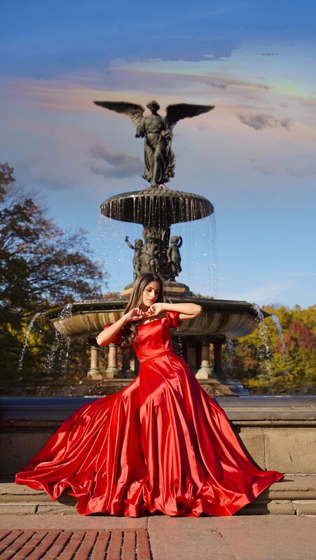 Roopsi Bansal in a red satin ballgown tracing a heart shape with her hands in front of the Bethesda Fountain angel statue in Central Park