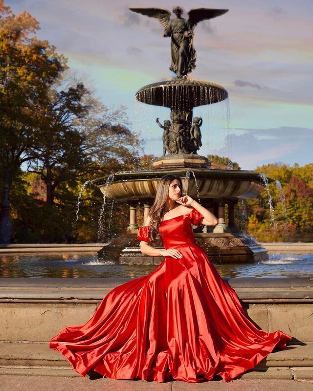 Roopsi Bansal in a red off-shoulder ballgown in front of the Bethesda Fountain angel statue in Central Park, Manhattan
