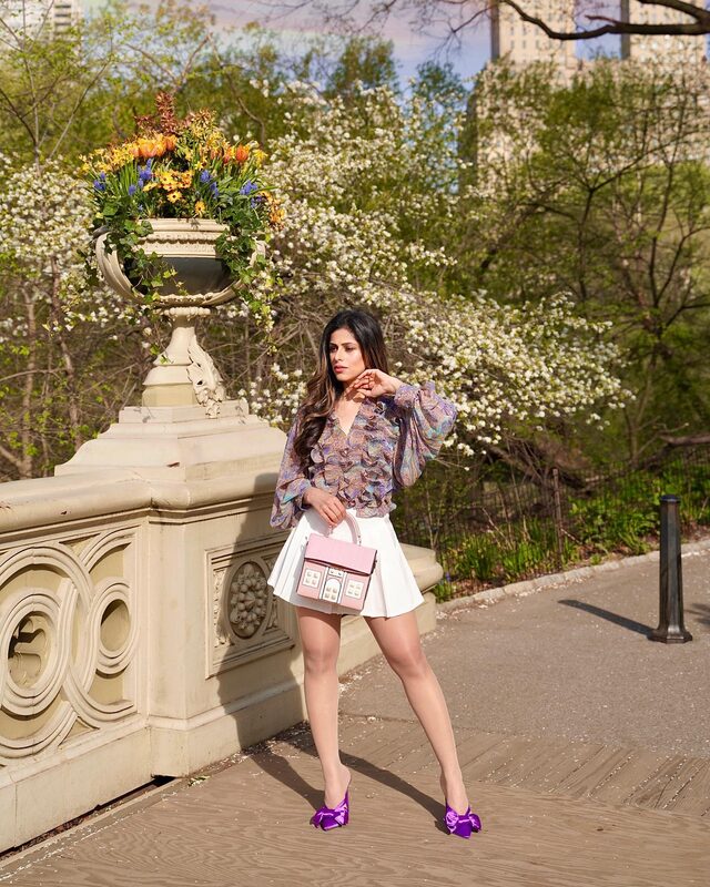 Roopsi Bansal in a ruffled paisley blouse and white pleated skirt standing by a stone balustrade with a flower-filled urn at Bethesda Terrace in Central Park