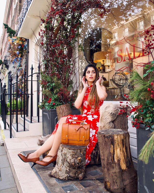 Roopsi Bansal in a red and white floral dress, seated on tree stumps outside a shop window decorated with berries and pinecones, holding a tan satchel bag