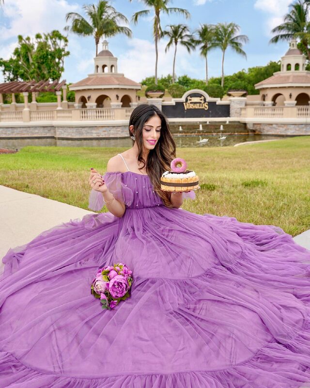 Roopsi Bansal in a sweeping lilac tulle off-shoulder gown, holding a small ladyfinger birthday cake topped with a pink donut, in front of the Versailles community gates in Wellington, Florida
