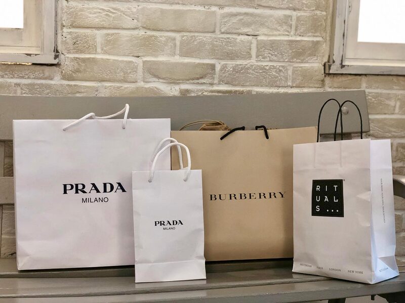 Shopping bags from Prada, Burberry, and Rituals lined up on a stone bench on Oxford Street, London