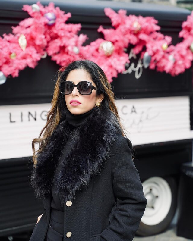 Roopsi Bansal in a black faux fur-trim coat and oversized sunglasses in front of the pink-decorated Lingua Franca café truck