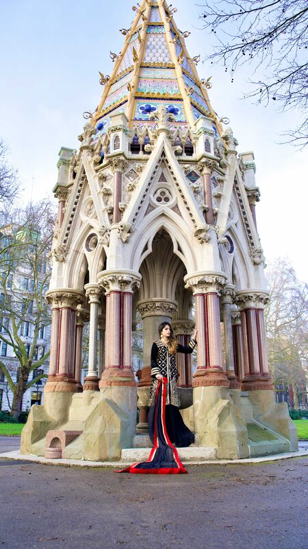 Roopsi Bansal in a black and gold embroidered long jacket with a red, black and cream striped train, standing inside an ornate Gothic stone memorial