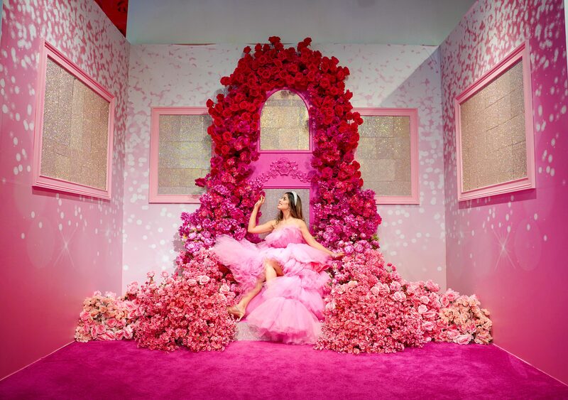 Roopsi Bansal in a pink tulle ruffle dress and tiara, seated inside a room-sized archway covered in red and pink roses at a floral installation in Aventura Mall