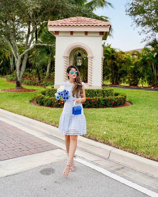 Roopsi Bansal in a white crochet dress and blue heart-shaped mirrored sunglasses, holding a bouquet of blue and white flowers and a small blue handbag, in front of a Florida community entrance archway