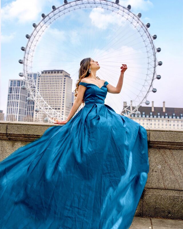 Roopsi Bansal in a teal off-shoulder ball gown, seated on a stone ledge with the London Eye rising directly behind her