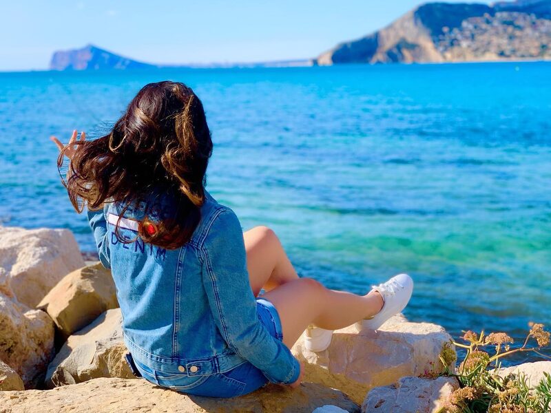 Sitting on rocks along the Spanish coast in a denim jacket, hair blowing in the wind, looking out at turquoise water and distant headlands