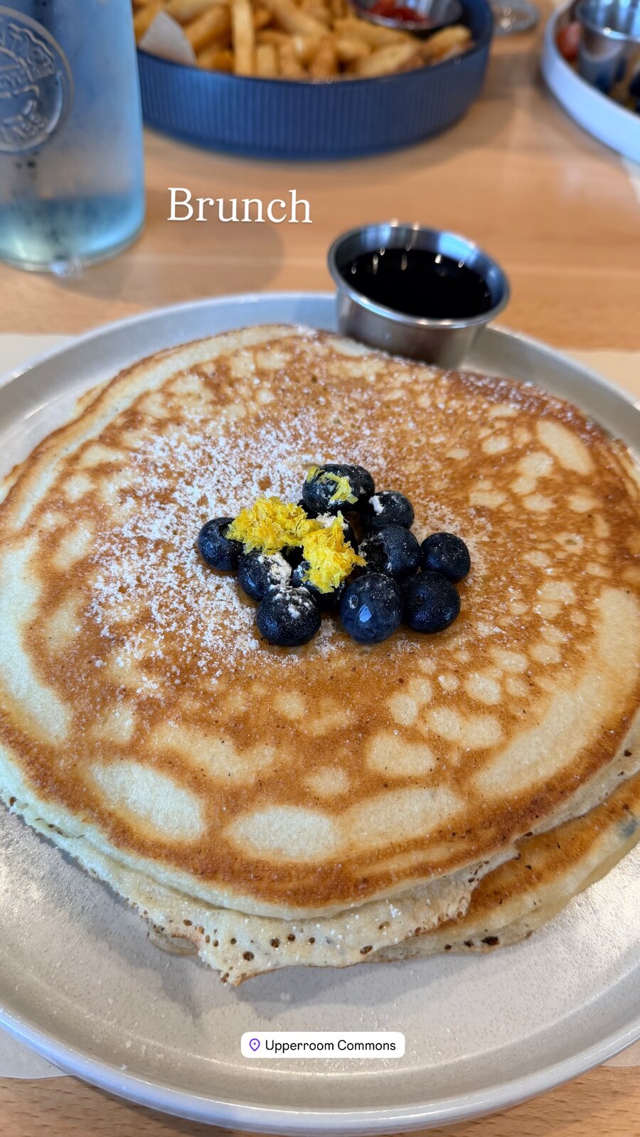 A tall stack of pancakes topped with blueberries, lemon zest, and powdered sugar, with a side of syrup, on-screen text reading Brunch, and a location tag for Upperroom Commons
