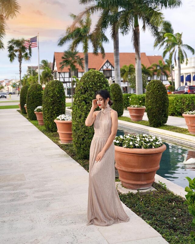 Roopsi Bansal in a metallic champagne gold halter gown from Adrianna Papell, standing beside a palm-lined canal with a swan swimming past, terracotta planters lining the walkway