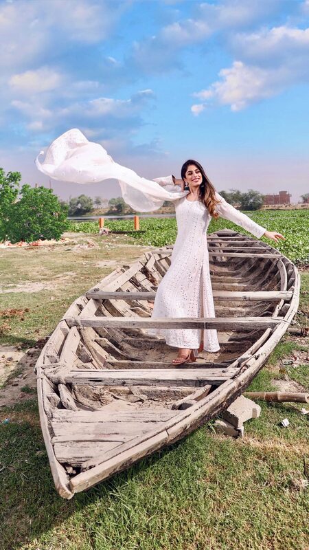 Roopsi Bansal in a white embroidered chikankari kurta, dupatta flying in the wind, standing in an old weathered wooden boat beside a field