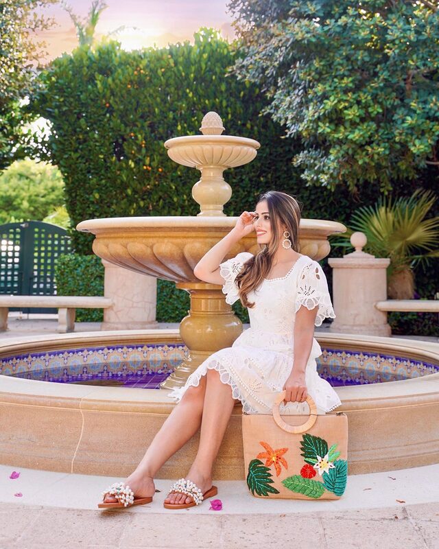 Roopsi Bansal in a white eyelet lace dress and pearl-embellished sandals, sitting beside a raffia tote embroidered with tropical flowers, in front of a tiled stone fountain