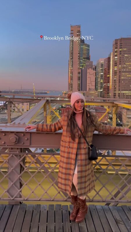 Roopsi Bansal in a plaid coat and pink beanie on the Brooklyn Bridge at sunset with the New York City skyline behind her