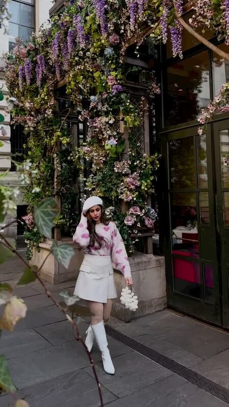Roopsi Bansal in a pink heart-print sweater, cream skirt, and beret standing in front of Bryant Park Grill's flower-covered facade