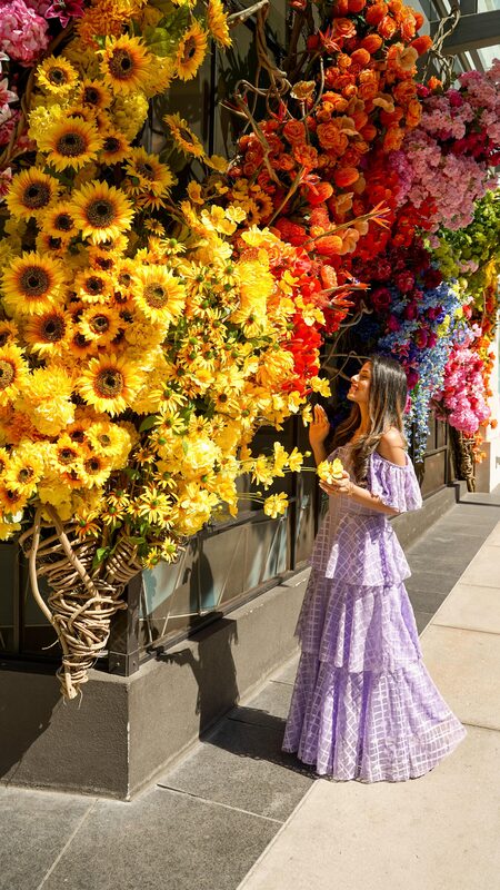Roopsi Bansal in a lilac off-shoulder ruffle dress from SHEIN, reaching toward a wall of sunflowers and orange roses in Buckhead, Georgia