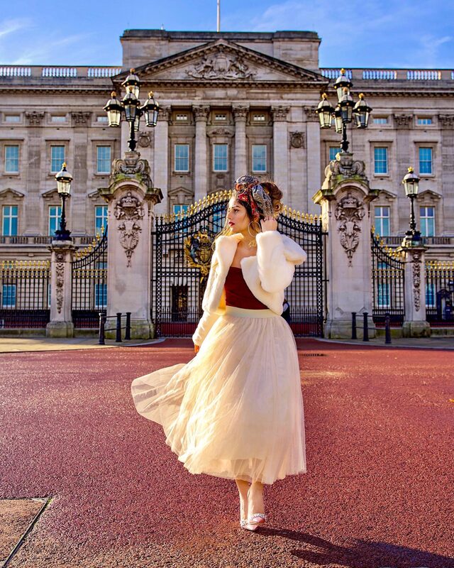 Roopsi Bansal in a cream tulle skirt, burgundy top, and cropped white faux-fur jacket, twirling in front of the gates of Buckingham Palace