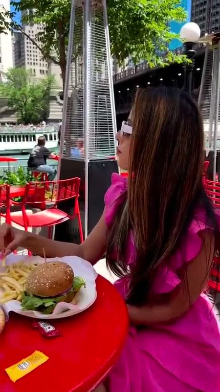 Roopsi Bansal in a pink ruffled dress at an outdoor table on the Chicago Riverwalk, reaching for a burger and fries
