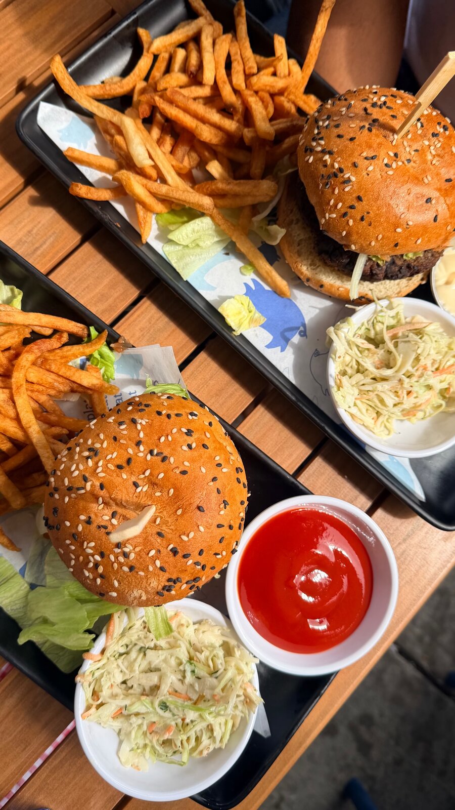 Two sesame-bun burgers with a pile of fries and coleslaw on a wooden picnic table