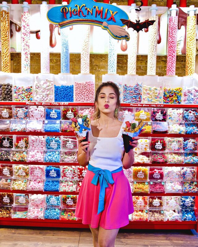 Roopsi Bansal in a white top and pink skirt with a blue bow belt, holding two cones of pick 'n' mix sweets in front of a wall of candy jars