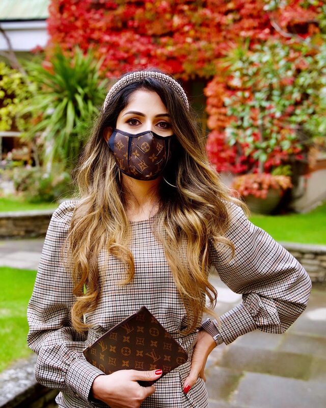 Roopsi Bansal in a checked long-sleeve top and jeweled headband, wearing a Louis Vuitton monogram face mask and holding a matching Louis Vuitton monogram clutch, in front of a red ivy wall