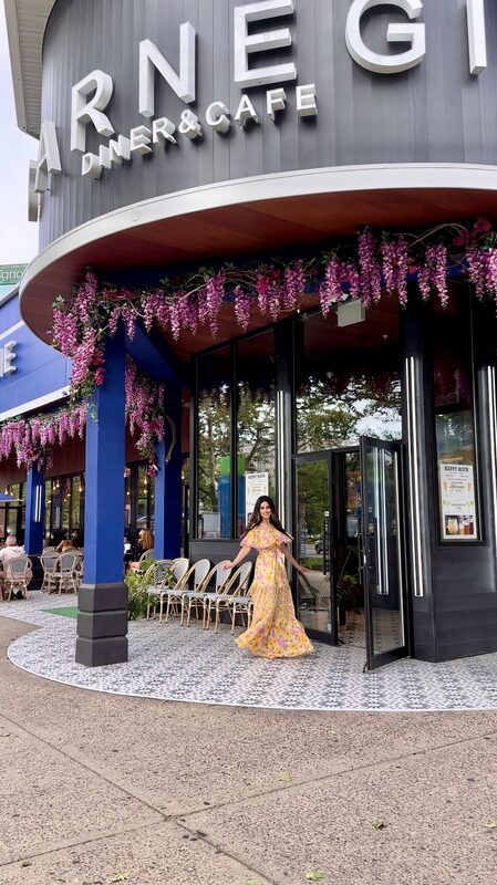 Roopsi Bansal in a yellow floral off-shoulder maxi dress standing outside Carnegie Diner's wisteria-draped entrance