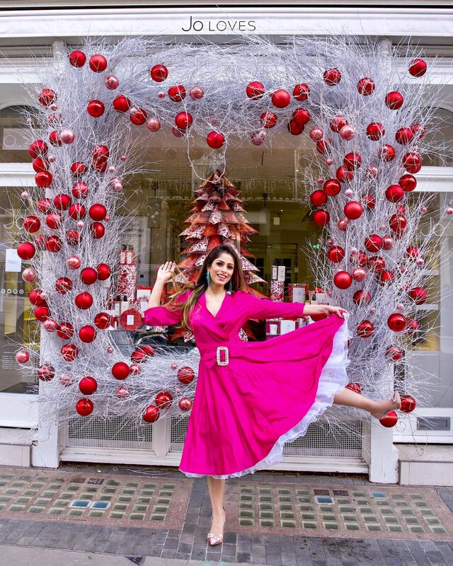 Roopsi Bansal in a hot-pink wrap dress, twirling in front of the Jo Loves storefront framed by a giant wreath of red Christmas baubles