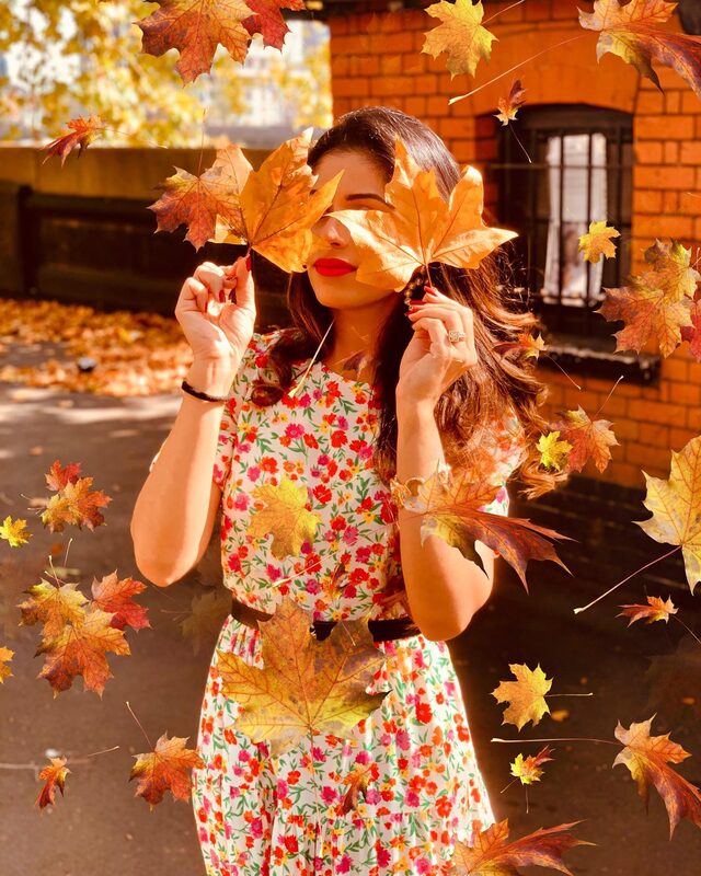Roopsi Bansal in a red and pink floral dress, holding an autumn leaf over one eye as more leaves fall around her in front of a brick building
