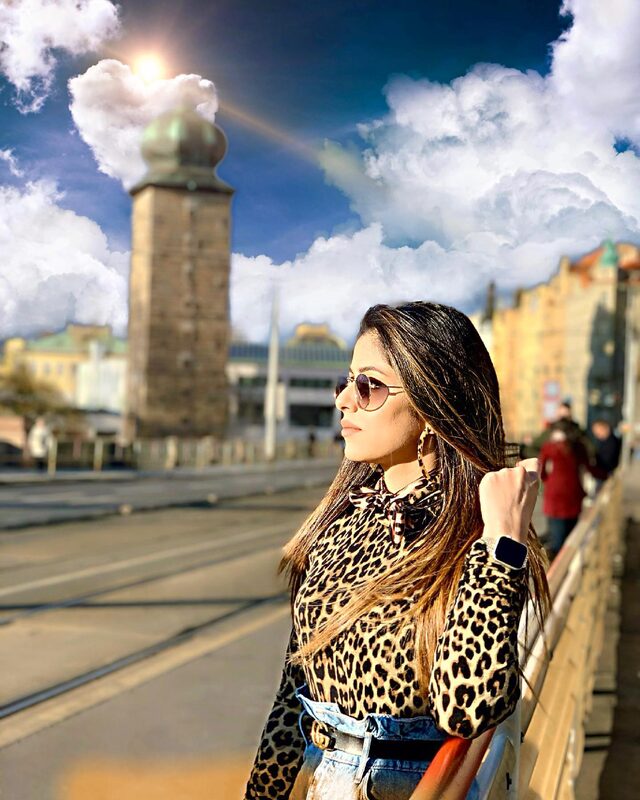 Standing on a bridge in Prague with a stone water tower behind and a dramatic sky overhead, wearing a leopard-print top, denim, and a Gucci belt