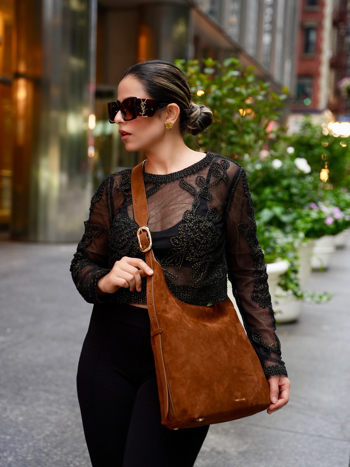 Roopsi Bansal walking a New York City street in a sheer black top, carrying the brown suede Aislin Hobo bag by Charles & Keith