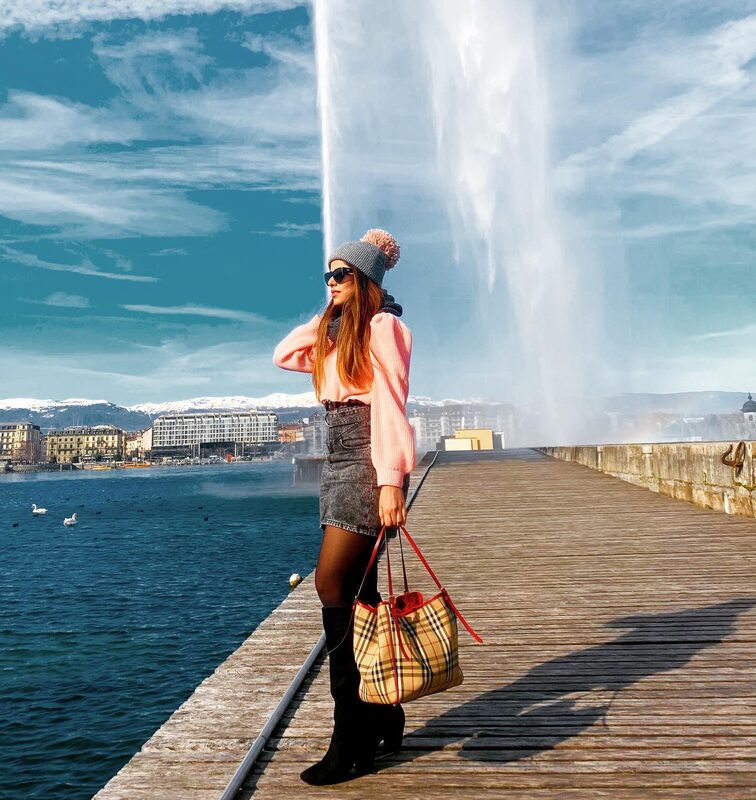 Standing on a wooden pier in Geneva, Switzerland, with the Jet d'Eau fountain spraying behind, wearing a pink sweater, grey pom-pom beanie, denim skirt, tall black boots, and carrying a Burberry check tote