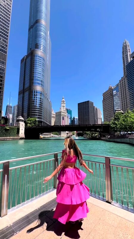Roopsi Bansal from behind in a hot pink cutout tiered dress, standing along the Chicago Riverwalk with Trump Tower rising behind her