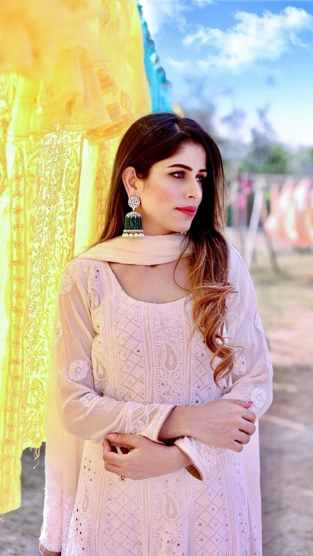 Roopsi Bansal in a white chikankari embroidered suit with a beige dupatta and emerald jhumka earrings, standing beside dupattas drying in the sun