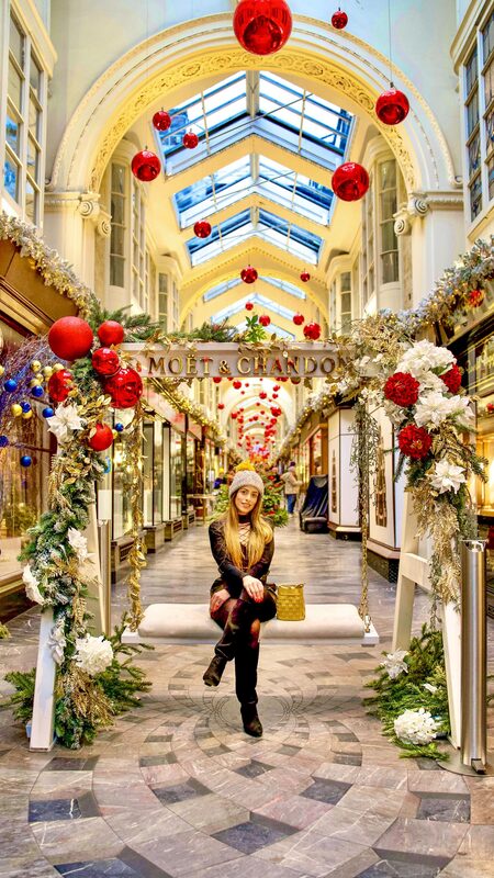 Roopsi Bansal in a plaid lace-up mini dress and knit beanie, seated on a swing bench under a Moet and Chandon sign in a Christmas-decorated arcade