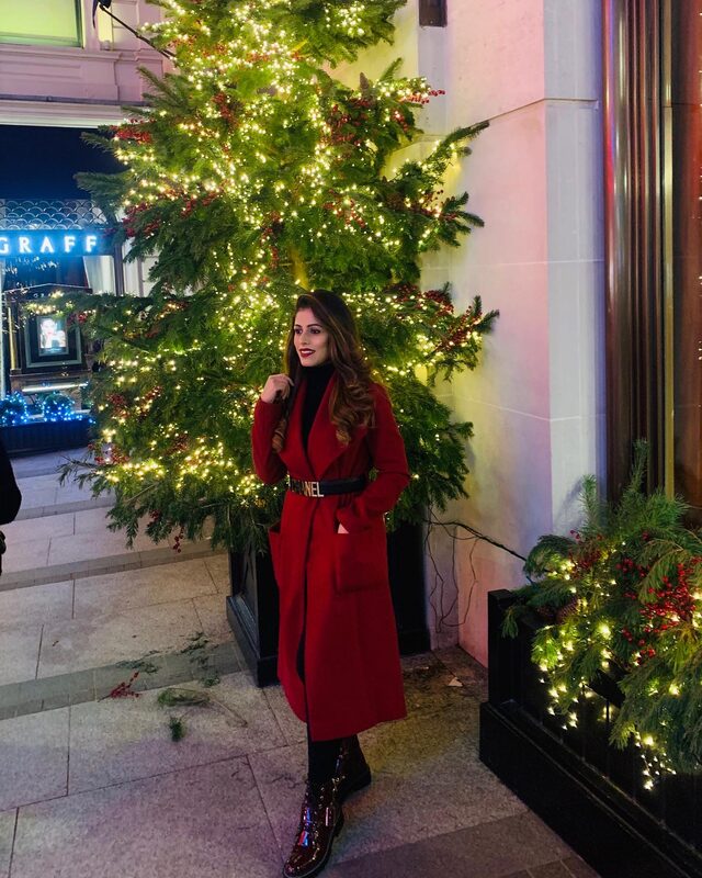 Standing in front of a fully lit Christmas tree outside a jewelry storefront in London, wearing a red wrap coat with a Chanel belt and patent boots