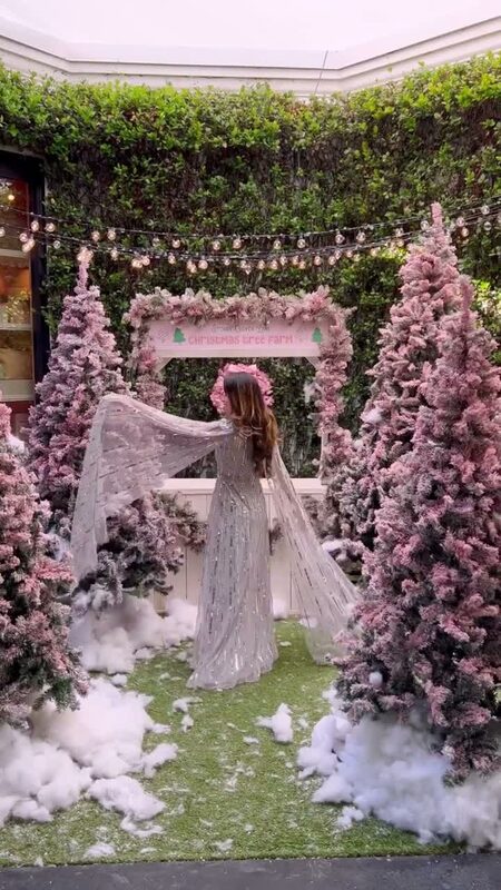 Roopsi Bansal in a silver sequined caped gown and flower crown, arms spread, standing among pink-flocked Christmas trees at a Christmas Tree Farm display
