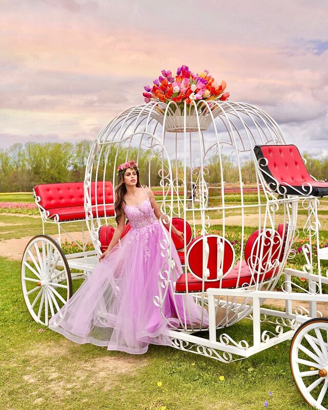 Roopsi Bansal in a lilac ballgown and flower crown standing beside a white Cinderella-style pumpkin carriage prop at Holland Ridge Farms