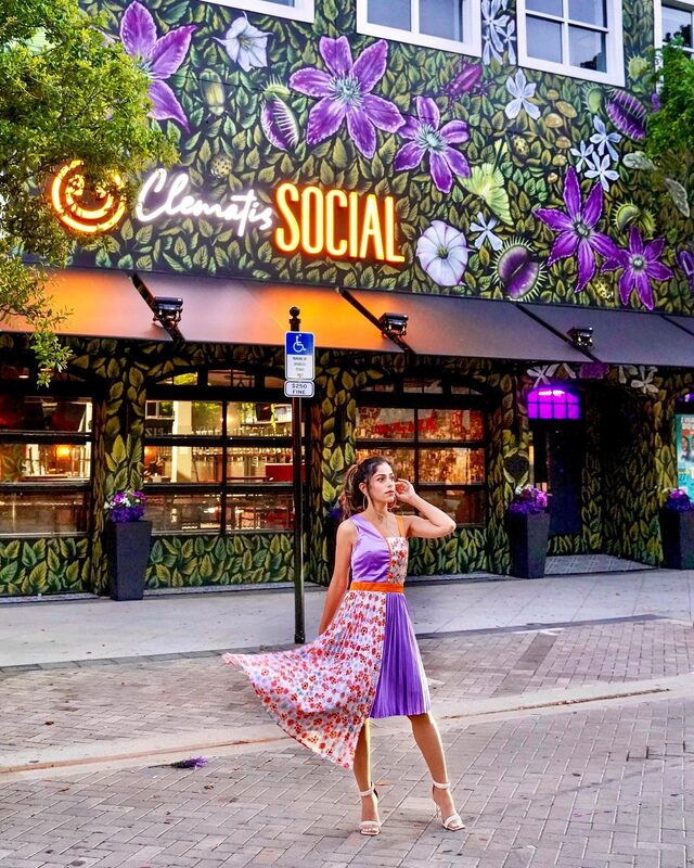 Roopsi Bansal in a purple and floral-print colorblock dress from SHEIN with orange trim, walking past the neon 'Clematis Social' sign and a mural of purple flowers