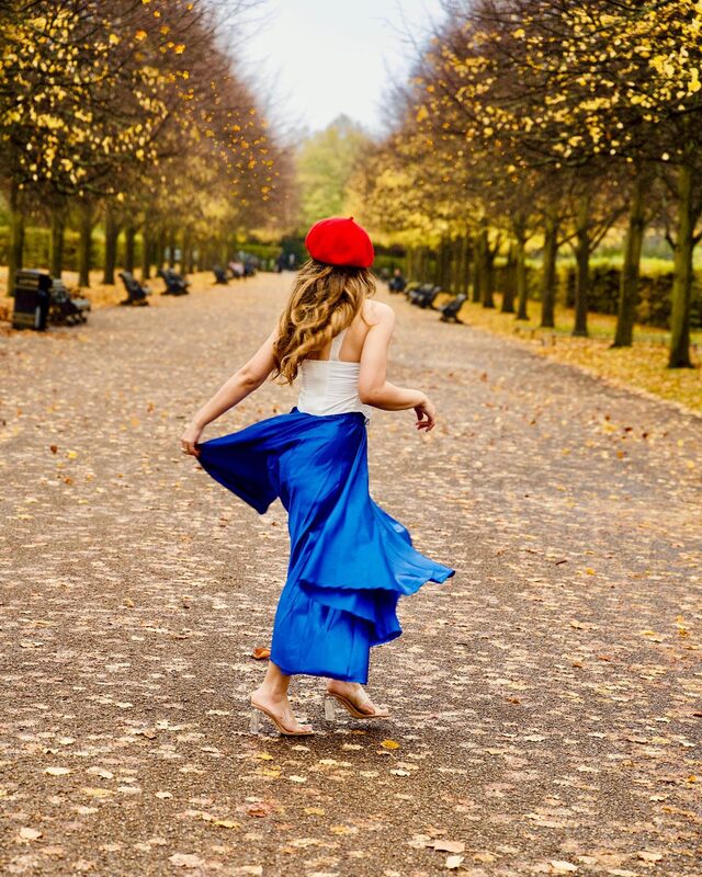 Roopsi Bansal in a red beret, white corset top and cobalt blue satin skirt, twirling down a tree-lined path in Regent's Park
