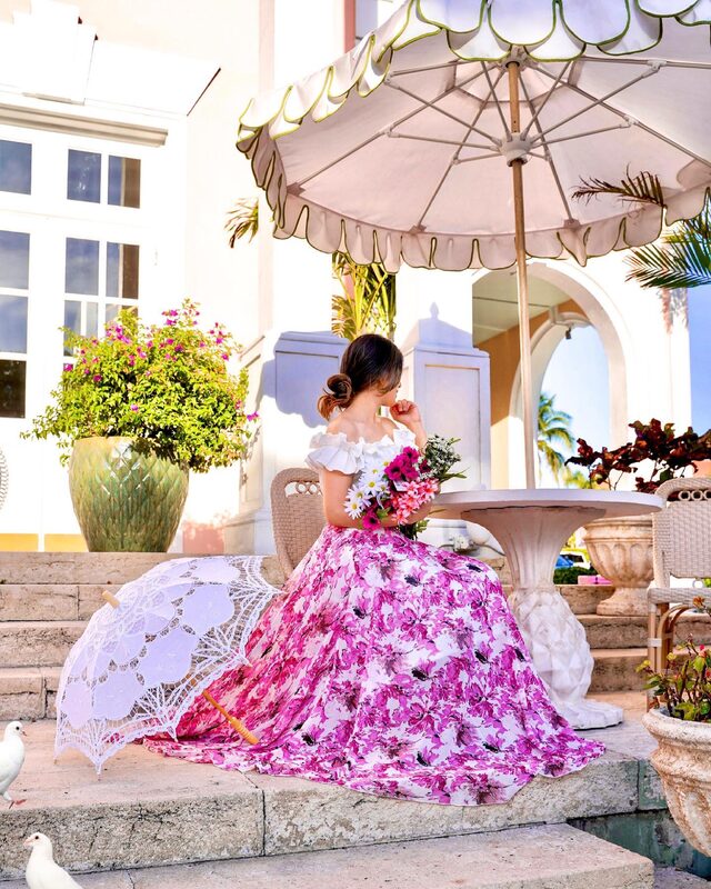 Roopsi Bansal in a magenta floral maxi skirt and white ruffle top, seated at a courtyard table with a lace parasol and white doves at her feet at the Colony Hotel, Palm Beach