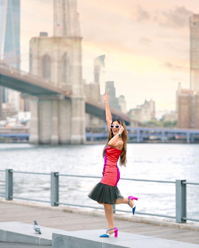 Roopsi Bansal in a red and pink colorblock dress with a black tulle hem, pointing at the sky in front of the Brooklyn Bridge