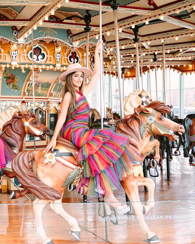 Roopsi Bansal in a rainbow tiered tulle dress sitting on a carved carousel horse at Jane's Carousel in Brooklyn