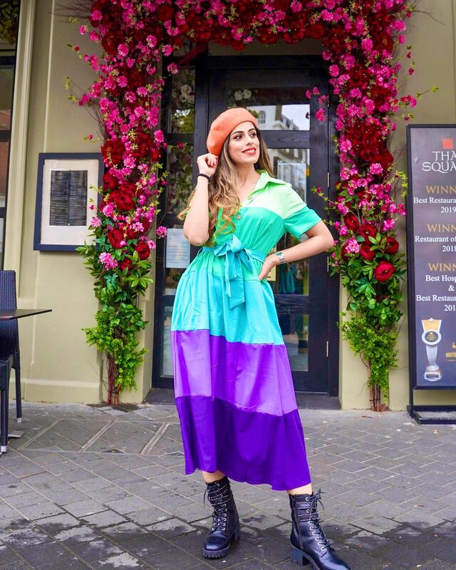Roopsi Bansal in a green-to-purple ombre midi dress, an orange beret, and combat boots, standing in front of a doorway framed by red roses