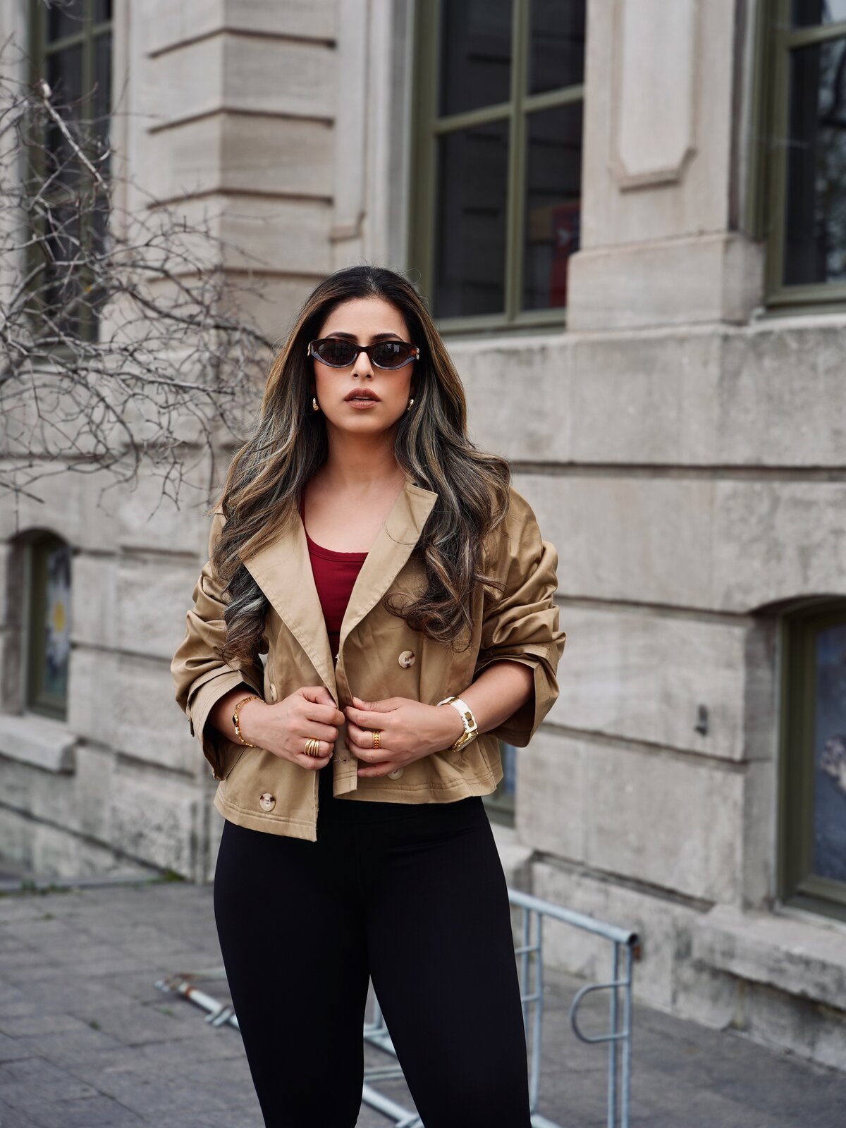 Roopsi Bansal in a cropped khaki trench coat over a red top, sunglasses and gold jewelry, standing in front of a stone building