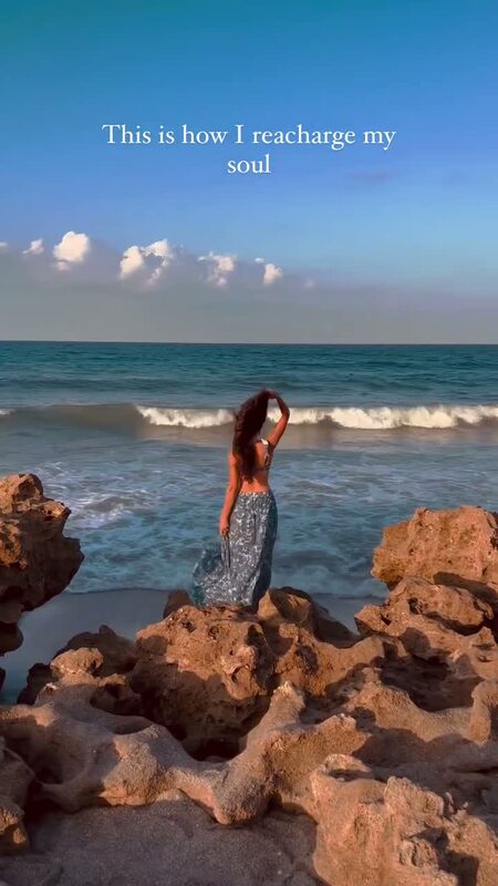 Roopsi Bansal standing on the rocky shoreline at Coral Cove Park, Florida, facing the ocean waves