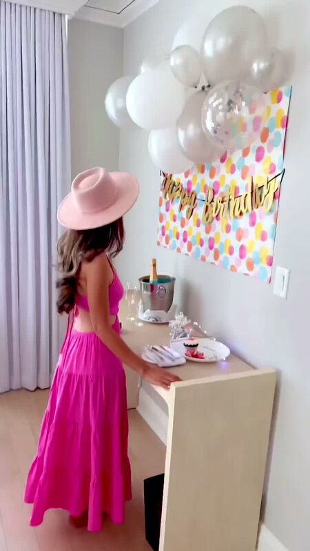 Roopsi Bansal in a hot pink crisscross backless tiered maxi dress and a pink boater hat, standing beside a table with a Happy Birthday banner, white balloons, and a champagne bucket