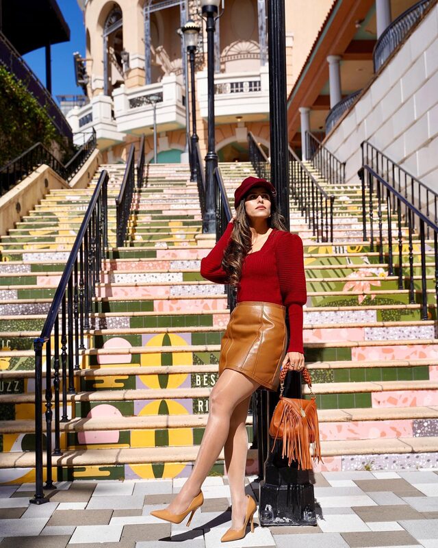Roopsi Bansal in a red puff-sleeve sweater, caramel faux-leather mini skirt, and burgundy cap, standing on colorful mural-painted steps in West Palm Beach