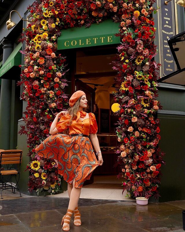 Roopsi Bansal in an orange puff-sleeve top and printed midi skirt with a Gucci GG logo belt and beret, standing in a floral archway outside a shop