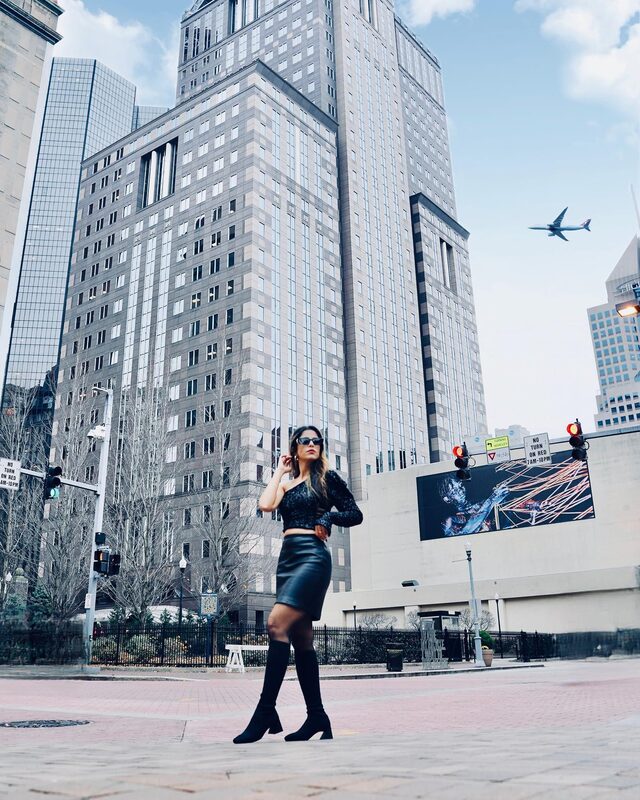 Roopsi Bansal in a black one-shoulder sequin top, leather skirt, and sunglasses walking across a downtown city street beneath tall office towers