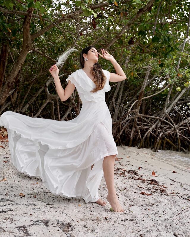 Roopsi Bansal barefoot in a white ruffle dress, holding a white feather, dancing among tangled mangrove roots on the sand at Coral Cove Park, Florida