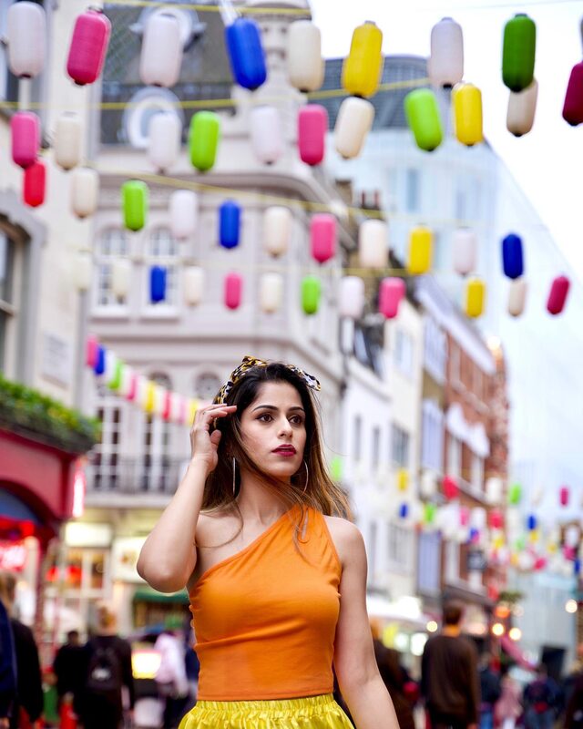 Roopsi Bansal in an orange one-shoulder top, yellow skirt and leopard-print headband, standing beneath a canopy of colorful paper lanterns on a London street